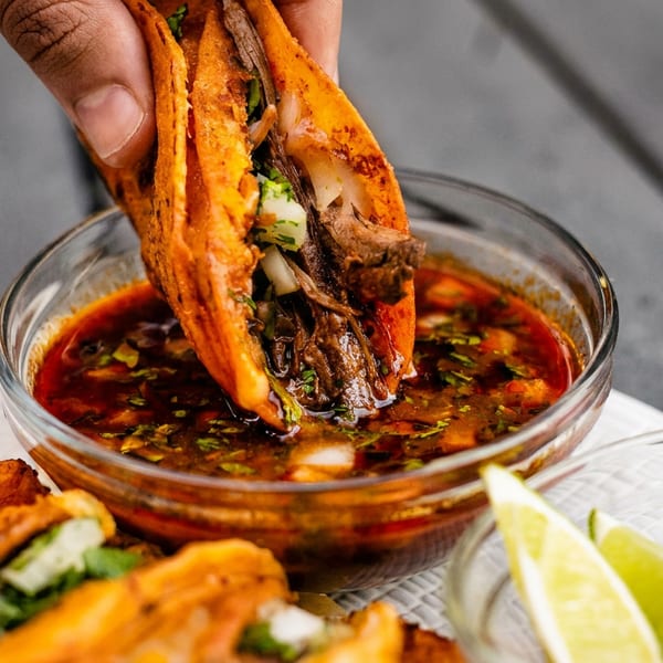 A hand dips a birria taco filled with meat, onions, and cilantro into a bowl of consommé.
