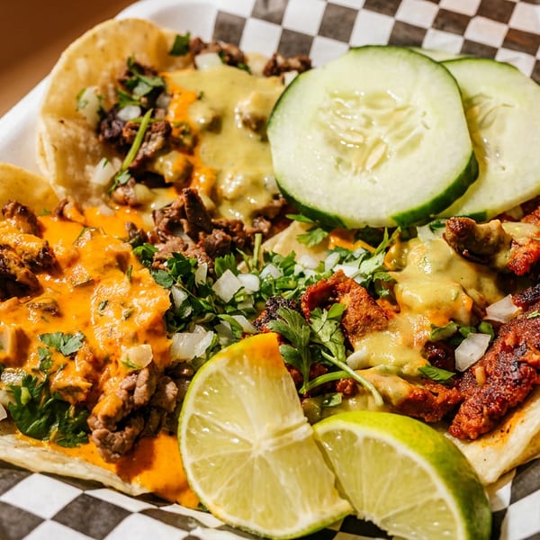 Three vibrant tacos with various meats, sauces, cilantro, onions, lime, and cucumber slices.