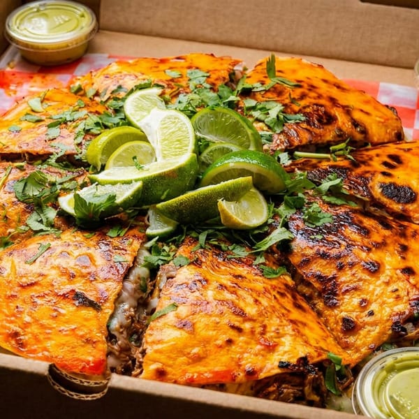Birria pizza, cut into slices, topped with cilantro and lime wedges, in a takeout box.