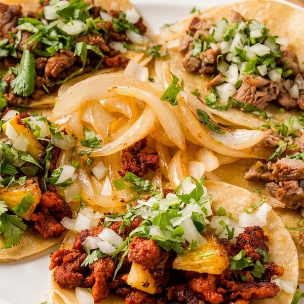 A white plate holds several tacos with various fillings, including meat, onions, and cilantro.